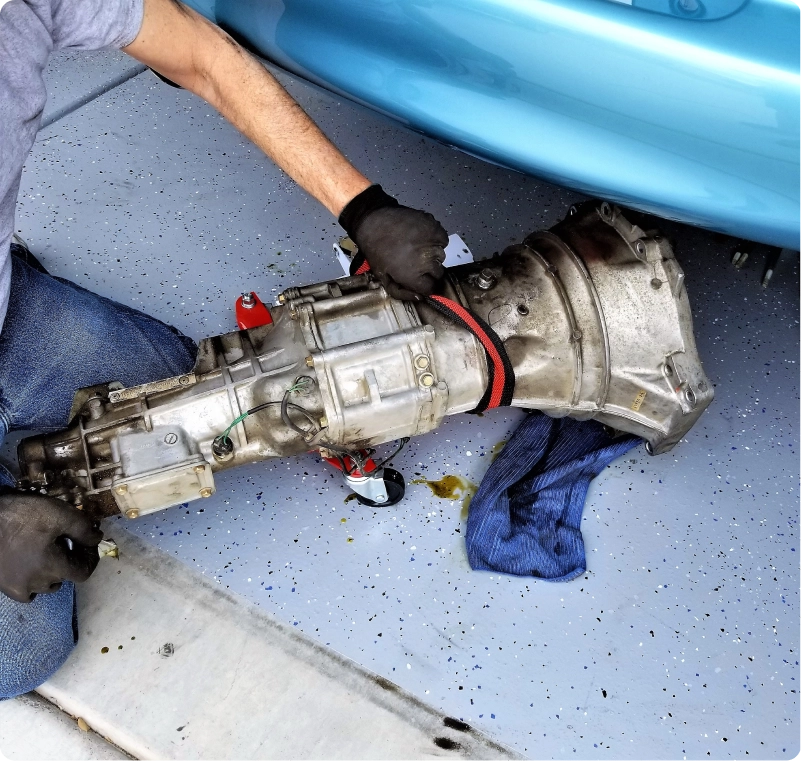 Mechanic handling a removed transmission beside a blue vehicle