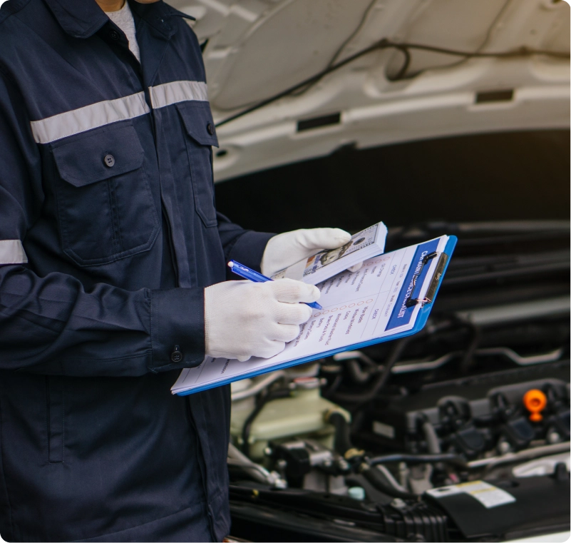 Mechanic wearing gloves recording inspection findings on a clipboard beside an open car hood